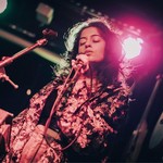 Artist aniqa dear sings onstage with a warm red light behind them and guitars on the wall. 
