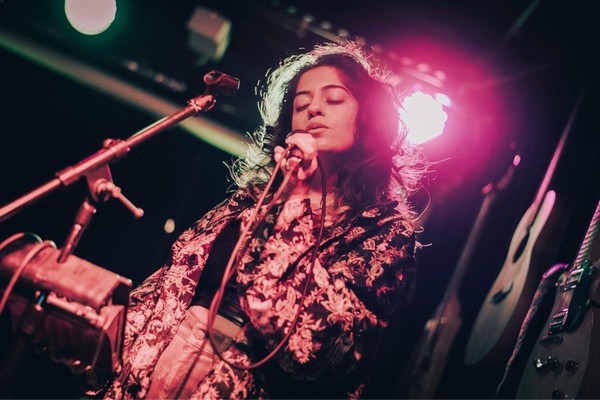 Artist aniqa dear sings onstage with a warm red light behind them and guitars on the wall. 