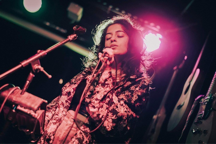 Artist aniqa dear sings onstage with a warm red light behind them and guitars on the wall. 