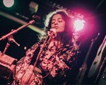Artist aniqa dear sings onstage with a warm red light behind them and guitars on the wall. 