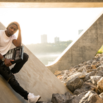 Rapper Arlo Maverick sits on a concrete slab under a bridge by the river with a drum pad machine in their hand. 