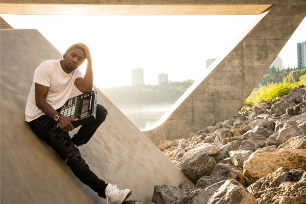 Rapper Arlo Maverick sits on a concrete slab under a bridge by the river with a drum pad machine in their hand. 