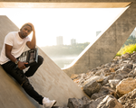 Rapper Arlo Maverick sits on a concrete slab under a bridge by the river with a drum pad machine in their hand. 