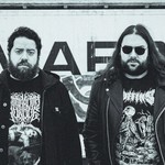 A black and white photo of duo Anthrodynia standing outside in front of a wall with graffiti. 