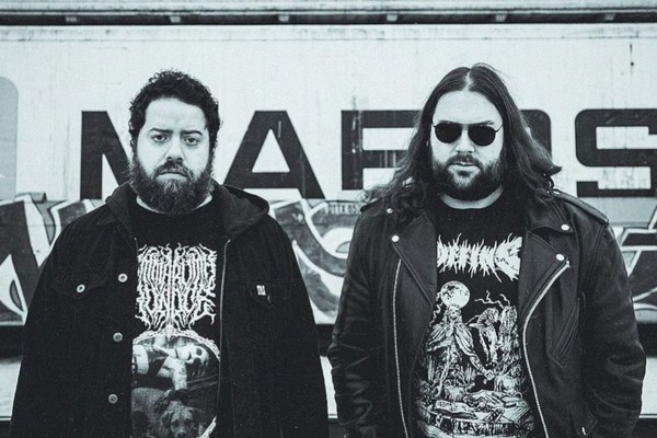 A black and white photo of duo Anthrodynia standing outside in front of a wall with graffiti. 