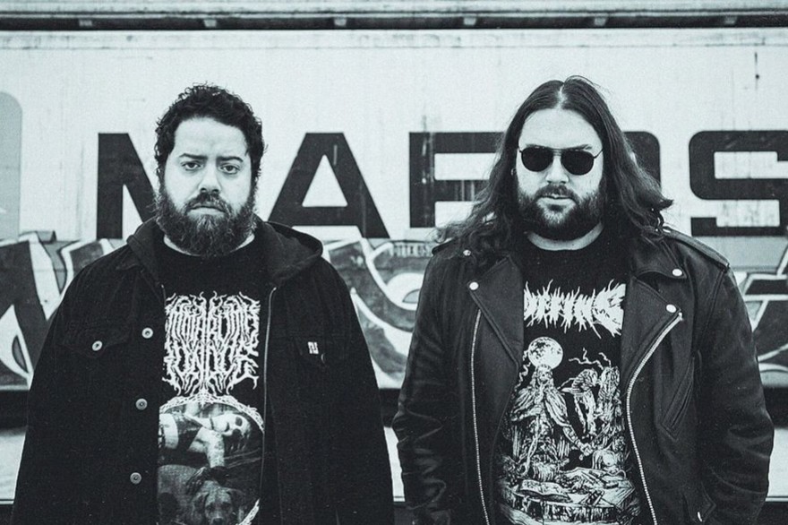 A black and white photo of duo Anthrodynia standing outside in front of a wall with graffiti. 