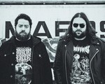 A black and white photo of duo Anthrodynia standing outside in front of a wall with graffiti. 