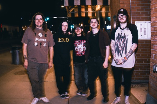 A colour photo of five-piece band Atrology Girl standing on a sidewalk outside of a brick building. 