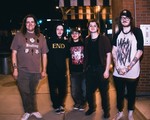 A colour photo of five-piece band Atrology Girl standing on a sidewalk outside of a brick building. 