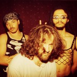 Three members of the group afk pose indoors for a photo. A member in the front has a white t-shirt and shaggy hair in their face. Two members stand in tank tops and pose straight faced in the background. 