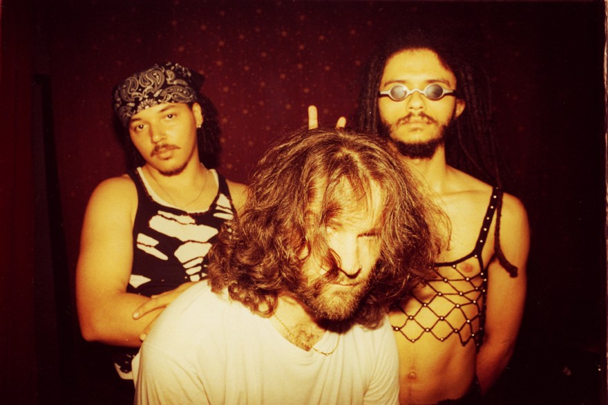Three members of the group afk pose indoors for a photo. A member in the front has a white t-shirt and shaggy hair in their face. Two members stand in tank tops and pose straight faced in the background. 