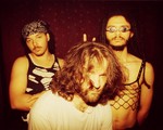 Three members of the group afk pose indoors for a photo. A member in the front has a white t-shirt and shaggy hair in their face. Two members stand in tank tops and pose straight faced in the background. 