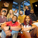 Eight-piece band (un)decided pose for a photo indoors with six members in the background sitting on a couch, and two members sitting on the floor in the foreground. 
