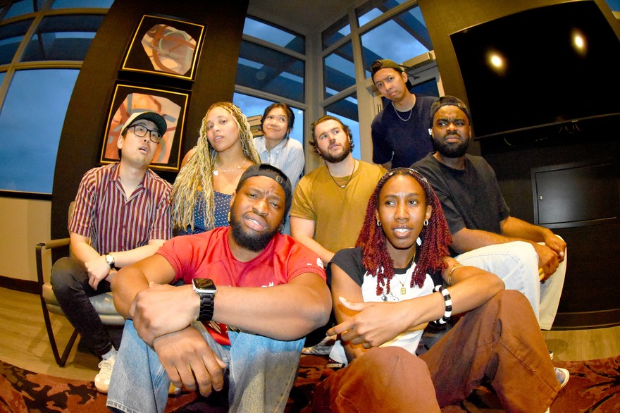 Eight-piece band (un)decided pose for a photo indoors with six members in the background sitting on a couch, and two members sitting on the floor in the foreground. 