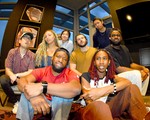 Eight-piece band (un)decided pose for a photo indoors with six members in the background sitting on a couch, and two members sitting on the floor in the foreground. 