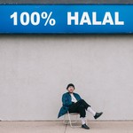 Artist Aladean Kheroufi sits on a lawn chair with a big concrete wall behind them and a sign on the top that says 100% Halal.  