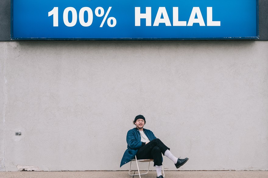 Artist Aladean Kheroufi sits on a lawn chair with a big concrete wall behind them and a sign on the top that says 100% Halal.  