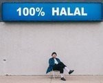 Artist Aladean Kheroufi sits on a lawn chair with a big concrete wall behind them and a sign on the top that says 100% Halal.  