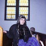 A colour photo of solo artist APACALDA sittiing in a pew in a chirch with a lavender wall behind them and a stain glass window. 