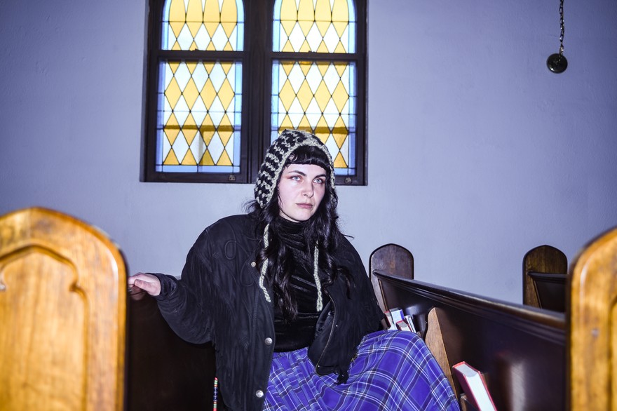 A colour photo of solo artist APACALDA sittiing in a pew in a chirch with a lavender wall behind them and a stain glass window. 