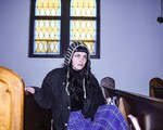 A colour photo of solo artist APACALDA sittiing in a pew in a chirch with a lavender wall behind them and a stain glass window. 