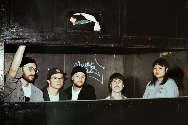 A colour photo of five-piece group Bad Bodies standing inside a cardboard compressor. They are seen from the neck and shoulders up.
