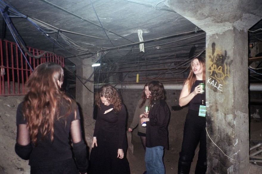 A candid colour photo shows five-piece group Babe Corner standing under a dingey concrete structure. Several members are holding cans of beer. 