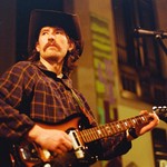 A grainy photo shows singer-songwriter Bennett Mitchell wearing a cowboy hat and playing guitar in an indoor venue. 