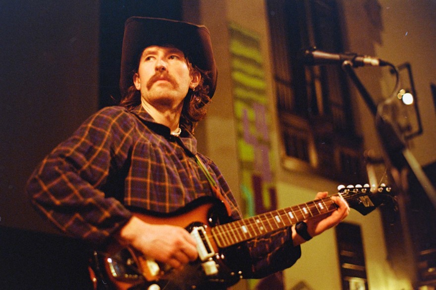 A grainy photo shows singer-songwriter Bennett Mitchell wearing a cowboy hat and playing guitar in an indoor venue. 