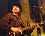 A grainy photo shows singer-songwriter Bennett Mitchell wearing a cowboy hat and playing guitar in an indoor venue. 