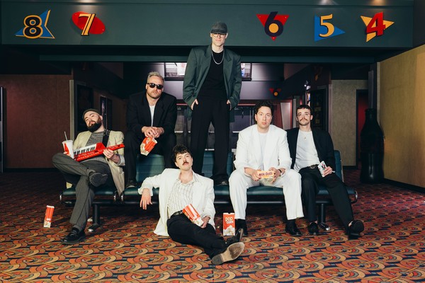 A colour photo shows six-piece band BETABOYS on a couch in the lobby of a movie theatre. Members hold popcorn and other snacks, and the member on the far left holds a keytar. 