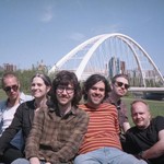 A grainy colour photo shows six-piece band BIG EVIL RAT sitting on the grass in front of a river with a big white arched bridge in the background. 