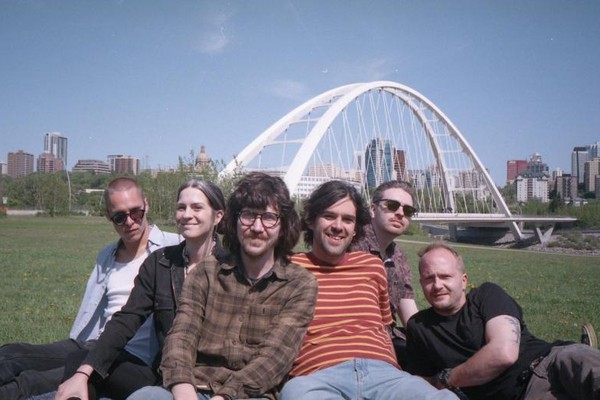 A grainy colour photo shows six-piece band BIG EVIL RAT sitting on the grass in front of a river with a big white arched bridge in the background. 