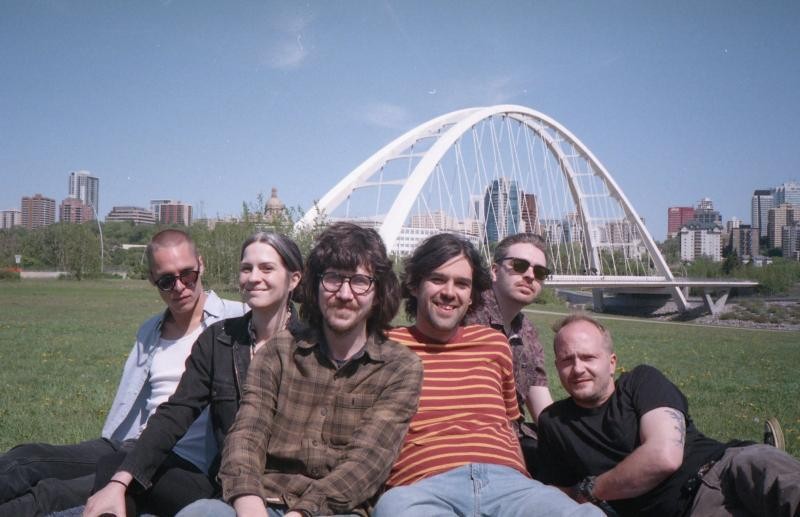 A grainy colour photo shows six-piece band BIG EVIL RAT sitting on the grass in front of a river with a big white arched bridge in the background. 