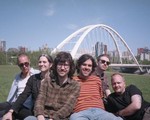 A grainy colour photo shows six-piece band BIG EVIL RAT sitting on the grass in front of a river with a big white arched bridge in the background. 