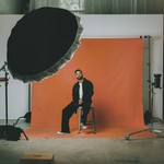 A colour photo shows artist Bijaan posing during a photo shoot. The sit on a stool in front of a rust orange backdrop with a large light facing them. 
