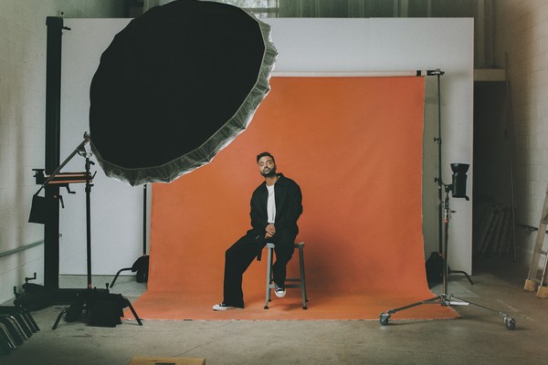 A colour photo shows artist Bijaan posing during a photo shoot. The sit on a stool in front of a rust orange backdrop with a large light facing them. 