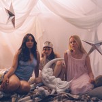 A colour photo shows three-piece Birds of Prrrey sitting on a bed with white draped sheets behind them, as well as plastic silver stars hanging, and white plastic moon sitting in front of them in the centre. 