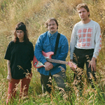 A colour photo shows three-piece Bluffing standing in a field with long green grass and wheat. The member in the middle holds a red guitar.
