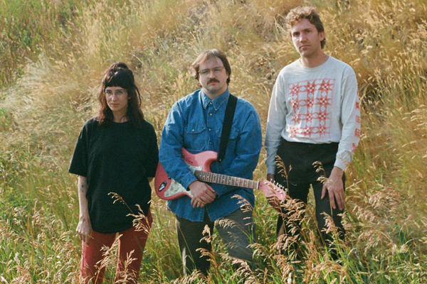A colour photo shows three-piece Bluffing standing in a field with long green grass and wheat. The member in the middle holds a red guitar.