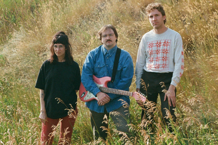 A colour photo shows three-piece Bluffing standing in a field with long green grass and wheat. The member in the middle holds a red guitar.