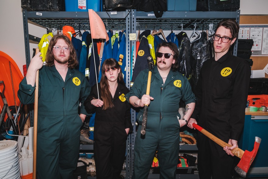 A colour photo shows four-piece band Brain Bent standing in a maintenace closet wearing matching jump suits, each of them holding tools like axes and sledgehammers. 