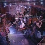 A blurry long exposure photo shows four-piece Brass Lip playing on stage with a white backdrop with their band name hanging behind them. 
