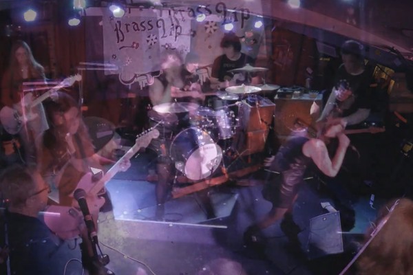 A blurry long exposure photo shows four-piece Brass Lip playing on stage with a white backdrop with their band name hanging behind them. 