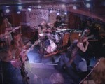 A blurry long exposure photo shows four-piece Brass Lip playing on stage with a white backdrop with their band name hanging behind them. 