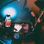 A colour photo taken from below shows four-piece BRIDGELAND playing on stage with blue and red lights shining on the members. 