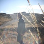 A grainy colour photo shows aritst Brock Geiger walking in a park with long strands of wheat in the foreground.