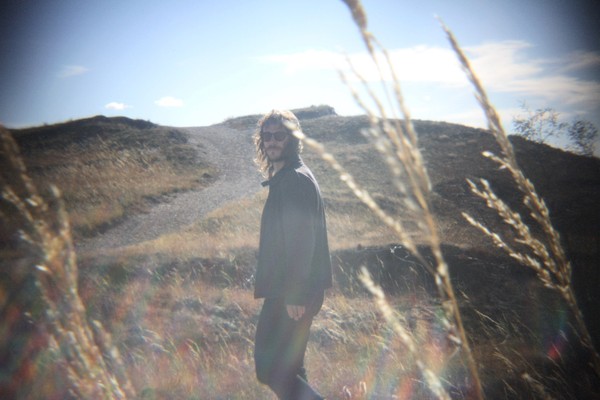 A grainy colour photo shows aritst Brock Geiger walking in a park with long strands of wheat in the foreground.