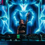 A vibrant colour photo shows Carissa Gem DJing in front of turntable with their left hand in the air and blue and red lightning-type projections behind them. 