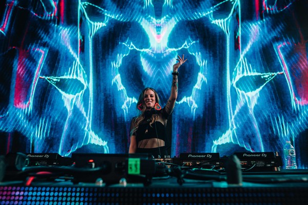 A vibrant colour photo shows Carissa Gem DJing in front of turntable with their left hand in the air and blue and red lightning-type projections behind them. 